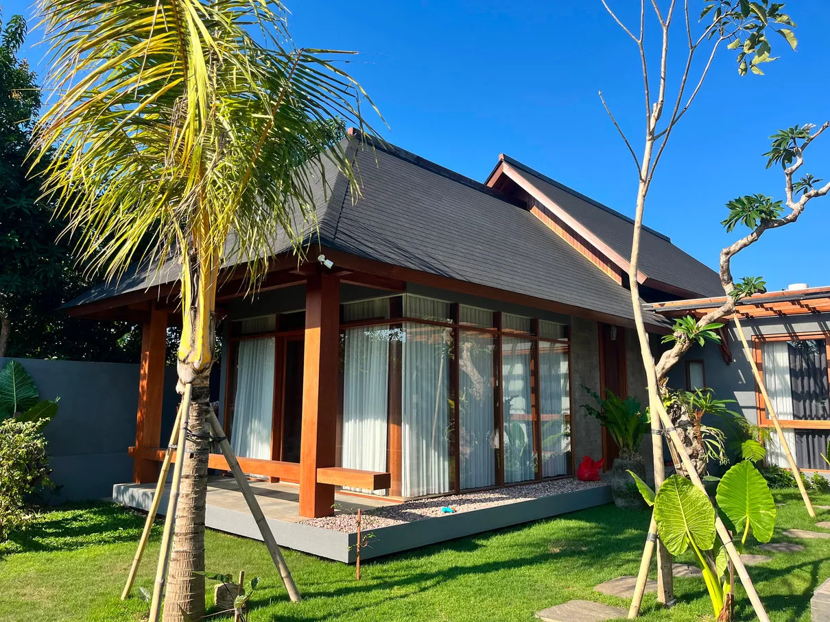 Rumah beratap miring dengan jendela kaca besar dan taman berpohon palem.