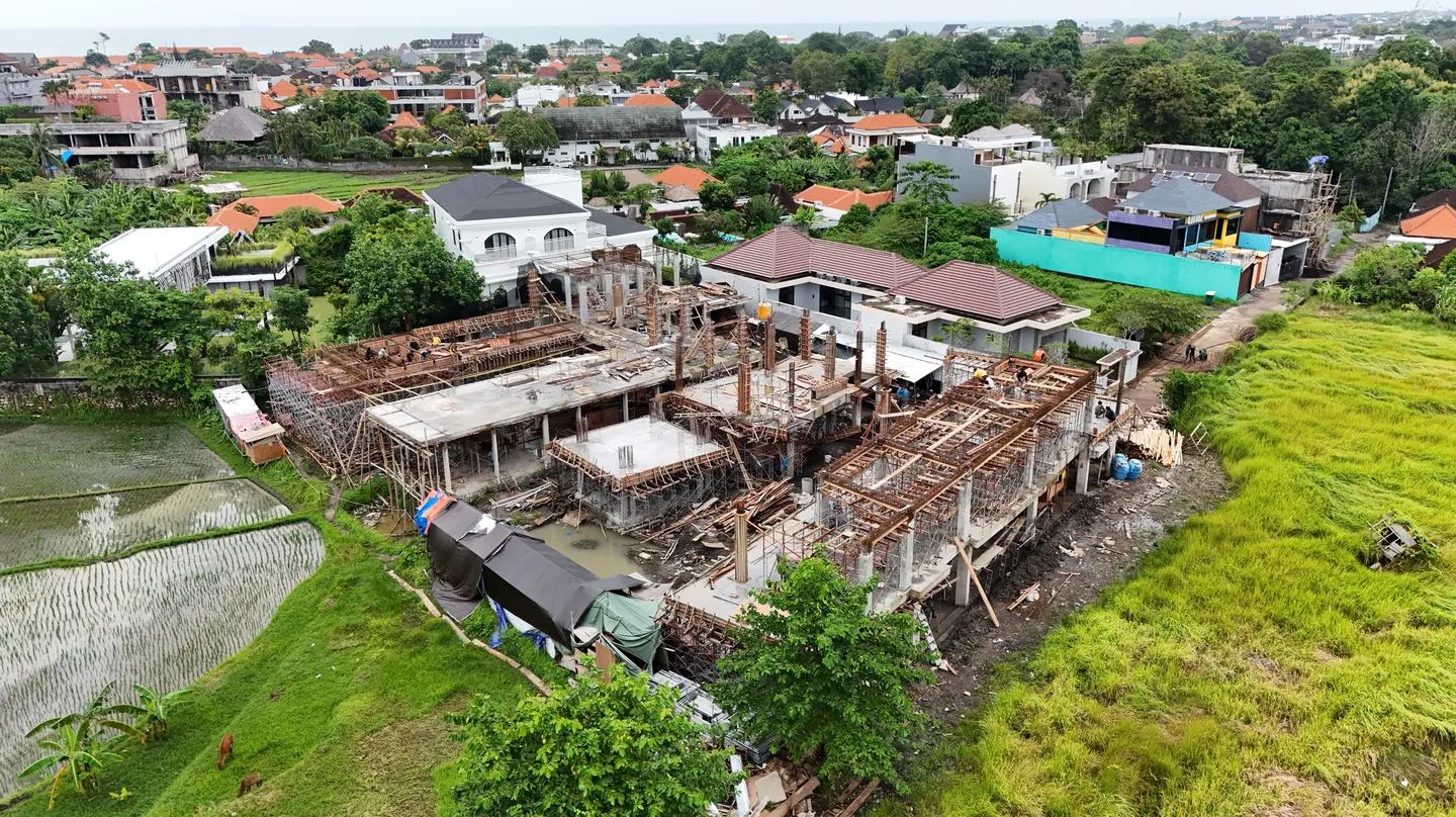 Foto udara bangunan villa dalam tahap konstruksi struktur beton dan perancah di antara sawah dan permukiman, hitam-putih.