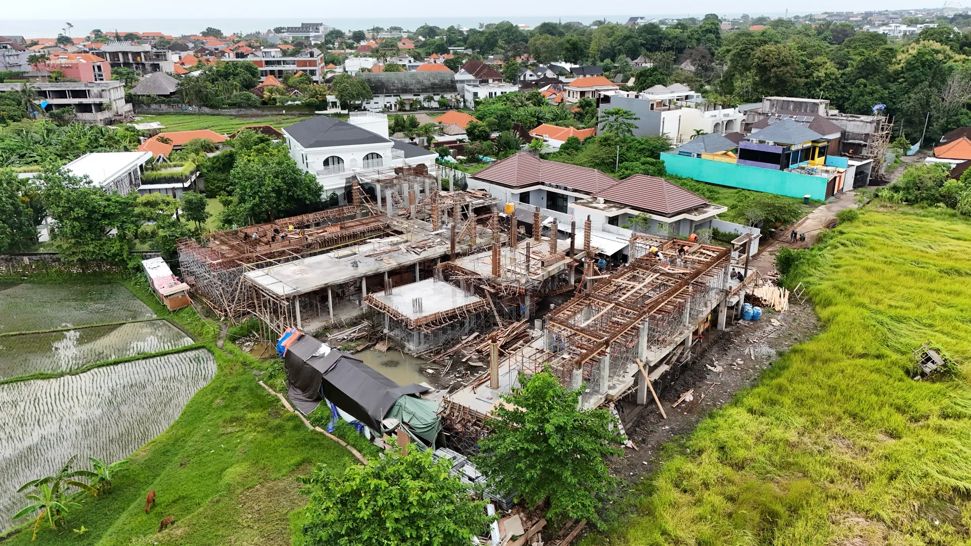 Foto udara bangunan villa dalam tahap konstruksi struktur beton dan perancah di antara sawah dan permukiman.