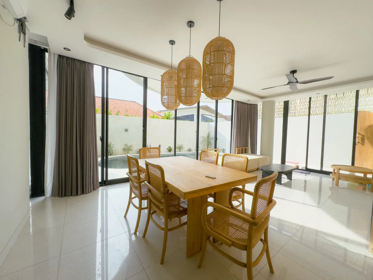 Ruang makan dengan meja kayu, lampu anyaman, dan jendela kaca besar.