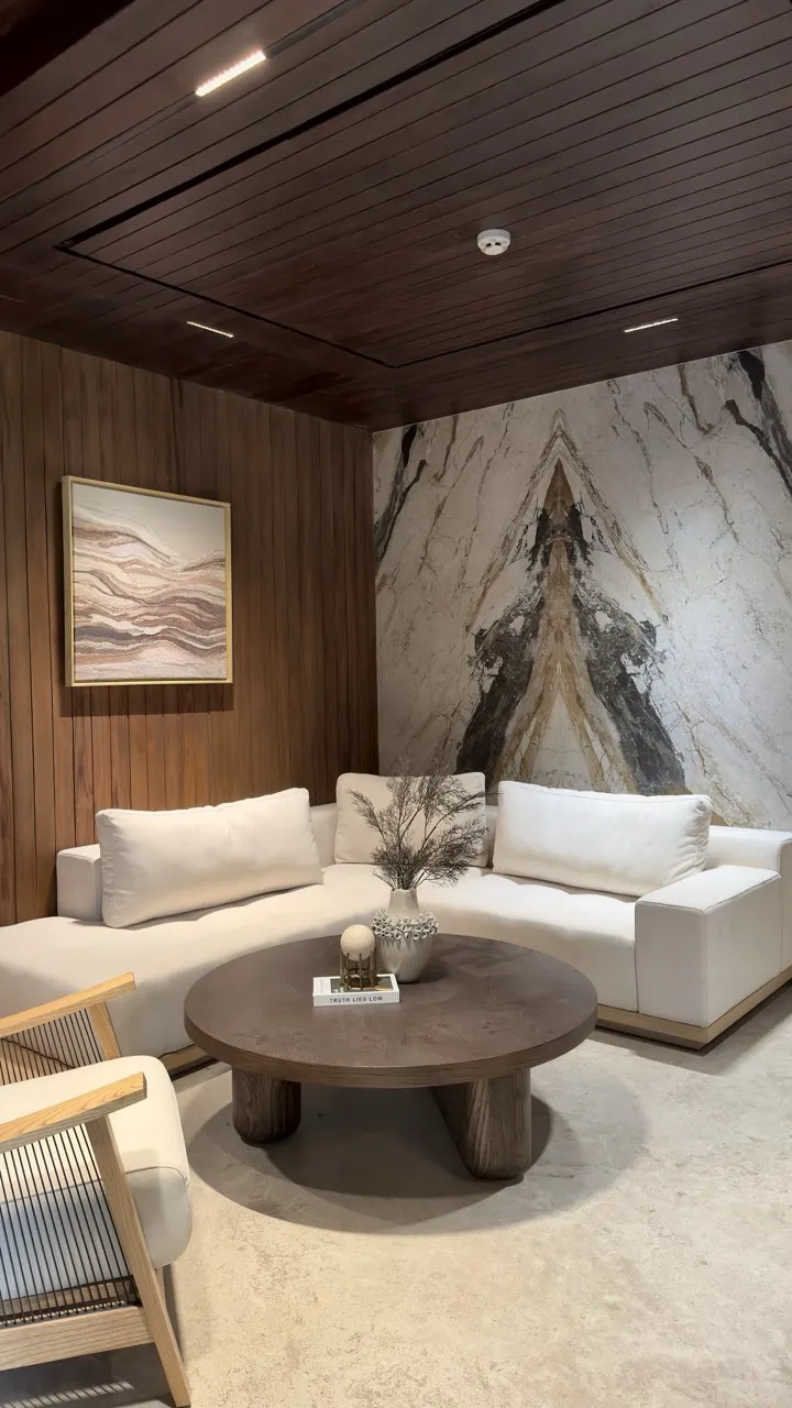 Lounge dengan dinding marmer bermotif dan sofa putih di bawah plafon kayu.